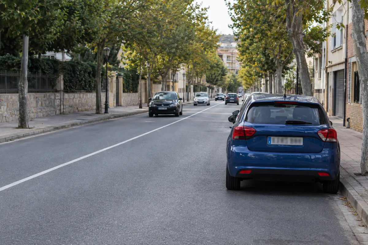 El coche aparcado a la derecha, la línea continua en el centro y la acera de enfrente más lejos.