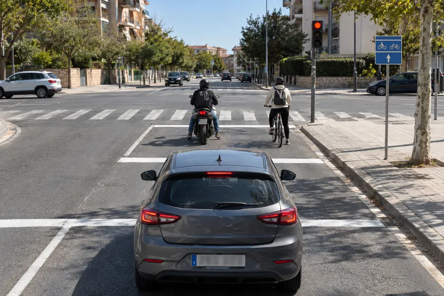 Un coche detenido ante un semáforo en rojo y, delante, una zona de espera marcada en el asfalto donde esperan una moto y una bicicleta