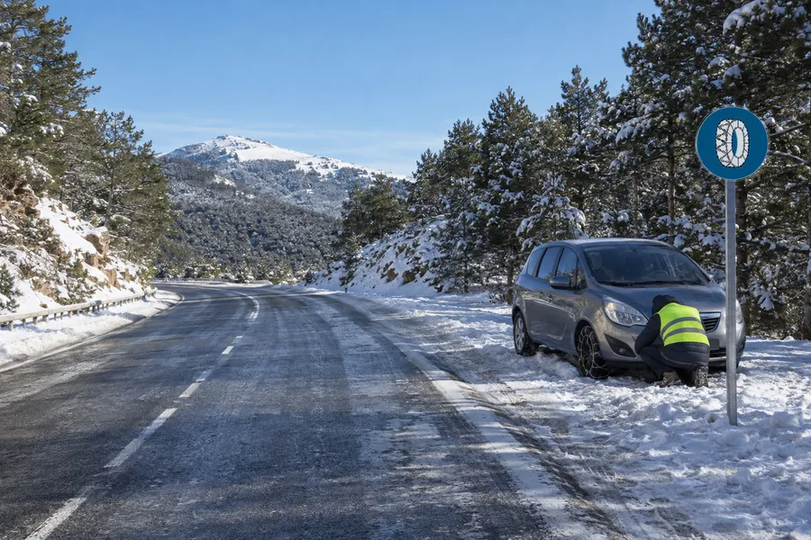 Un coche parado en una carretera nevada junto a la señal de cadenas para nieve, mientras una persona coloca las cadenas en una rueda
