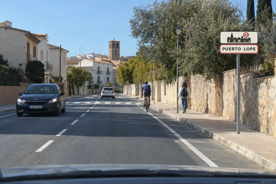 Una carretera a la entrada de un pueblo, con la señal de entrada a poblado junto a la calzada y un peatón y un ciclista más adelante