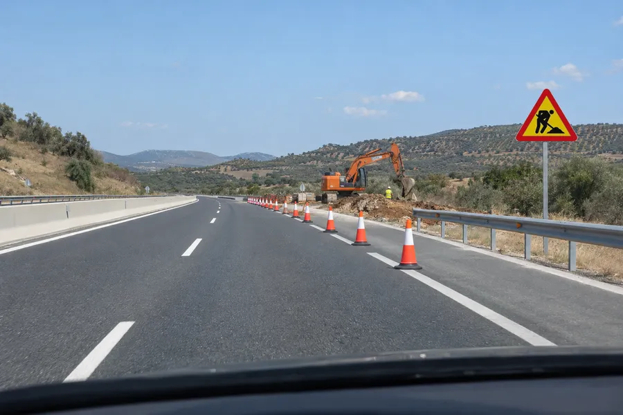 Un tramo de carretera en obras: la señal de obras lo anuncia, una fila de conos cierra el carril derecho y detrás hay una excavadora