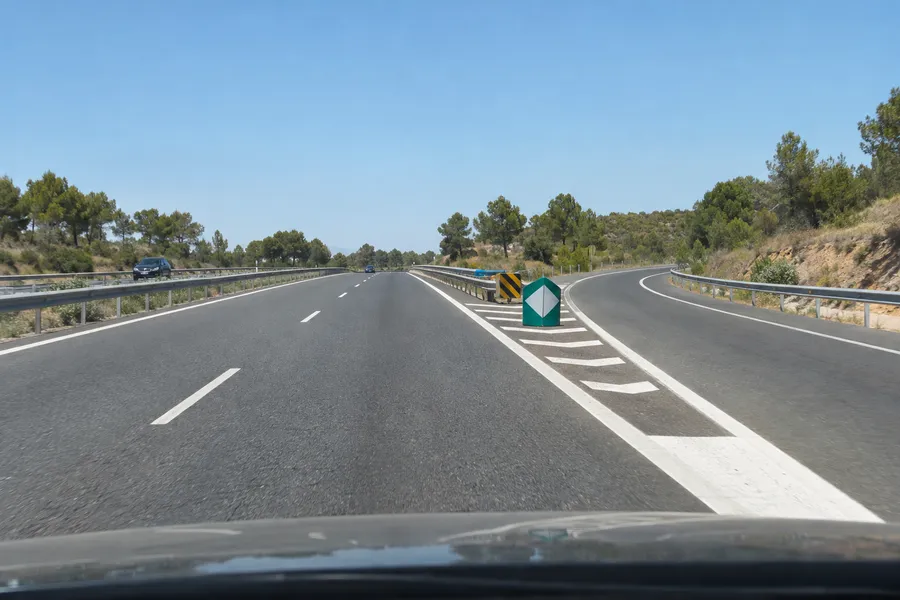 Una salida de autovía de día: un hito de vértice marca dónde se separan los carriles, con un atenuador de impacto delante y barreras de seguridad en el margen y la mediana