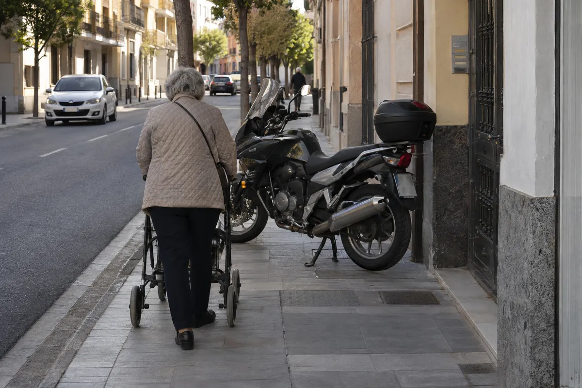 Una moto atraviesa la acera y una persona con andador tiene que detenerse.